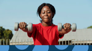 A woman putting on a red top holding 2kg weight in each hand in front of her chest.